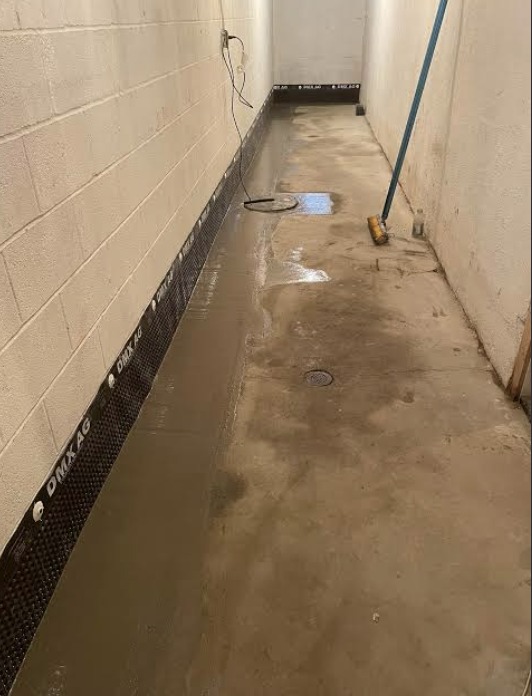 Basement hallway with dimpled DMX AG drainage membrane along the wall base, floor drain and wet concrete being squeegeed