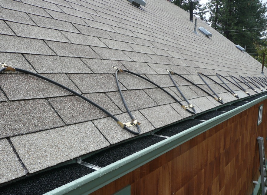 De-icing heating cable clipped in loops along the edge of a grey shingle roof above a green eavestrough