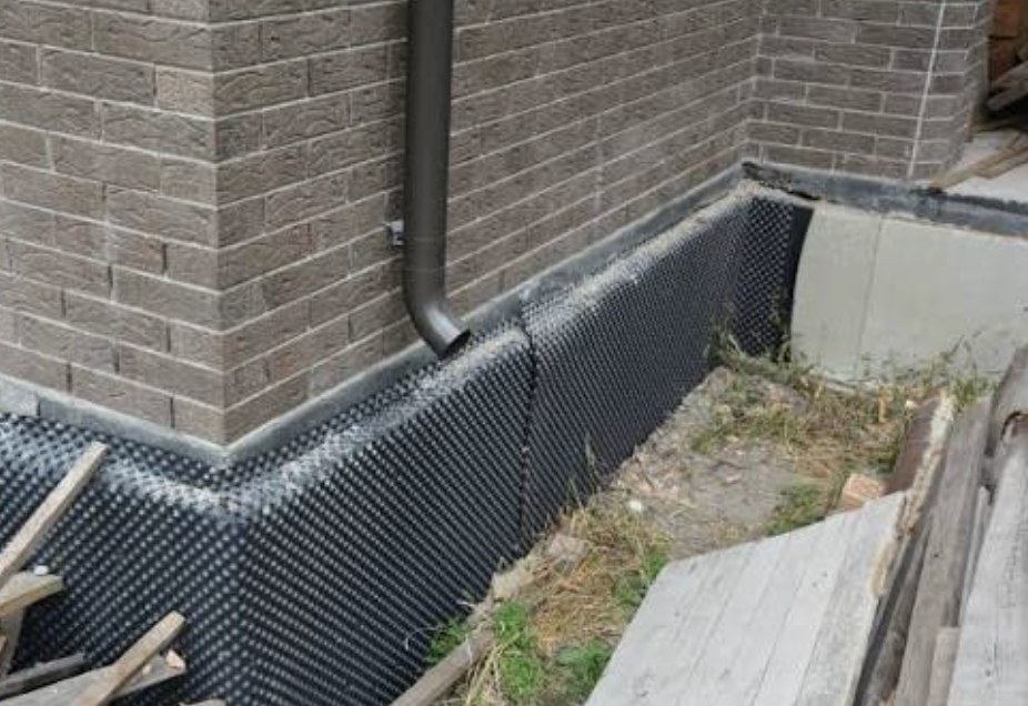 Dimpled damp-proofing membrane on a basement foundation wall beside a brick home and downpipe